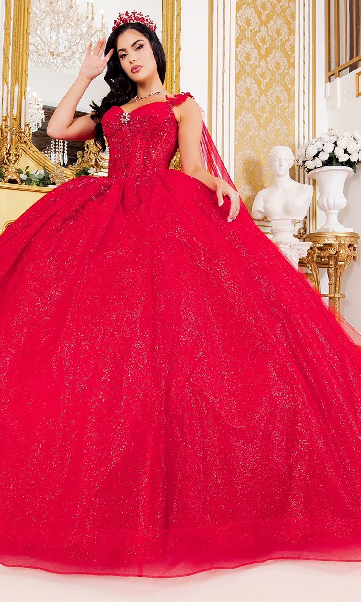 A woman wearing the Quinceanera Dress 8270J by Cinderella Couture in sparkling red stands in an ornate room adorned with gold decor, a chandelier, and a white bust statue in the background. The dress showcases glitter tulle and intricate 3D floral appliques that catch the light beautifully.